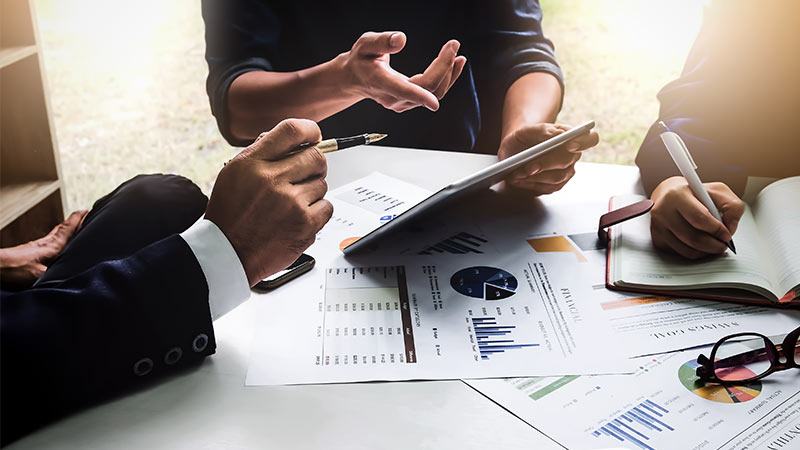 Three people sitting around a table working on a tablet and writing notes