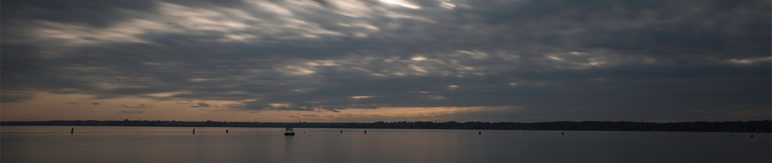 Orange sunset overcast by grey clouds, above a body of water.